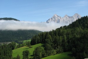 Man sieht ein Foto mit Berg, Wald und Wiesen. Es ist ein schönr Tag, in der Ferne ist Nebel zu erkennen.